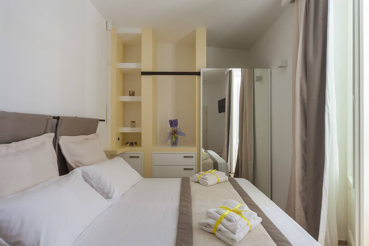 A hotel room with a bed, a wardrobe, a vanity with a mirror, and a small table with a vase of flowers.