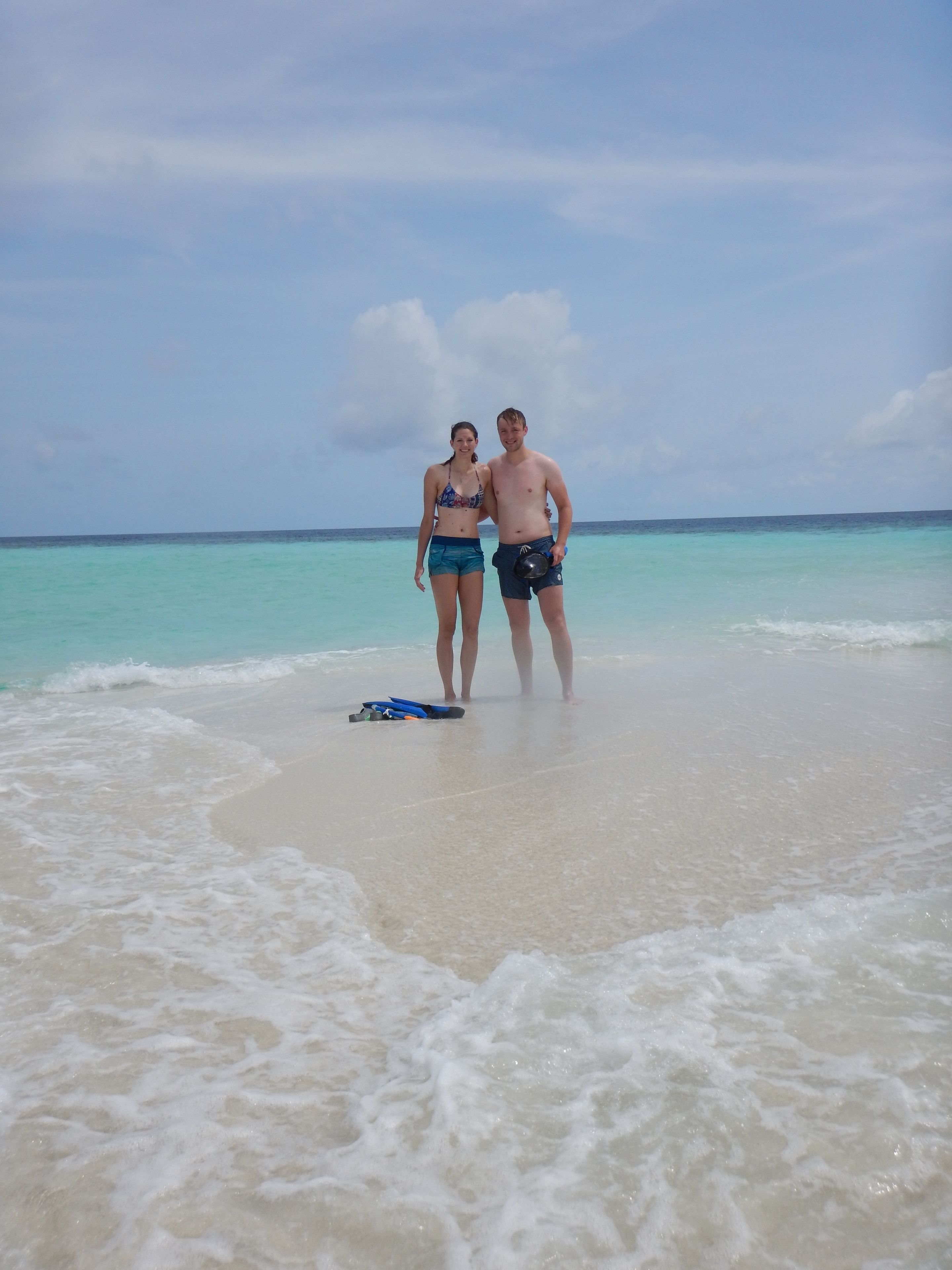 Two people standing on a beach with clear turquoise water and a calm sea.