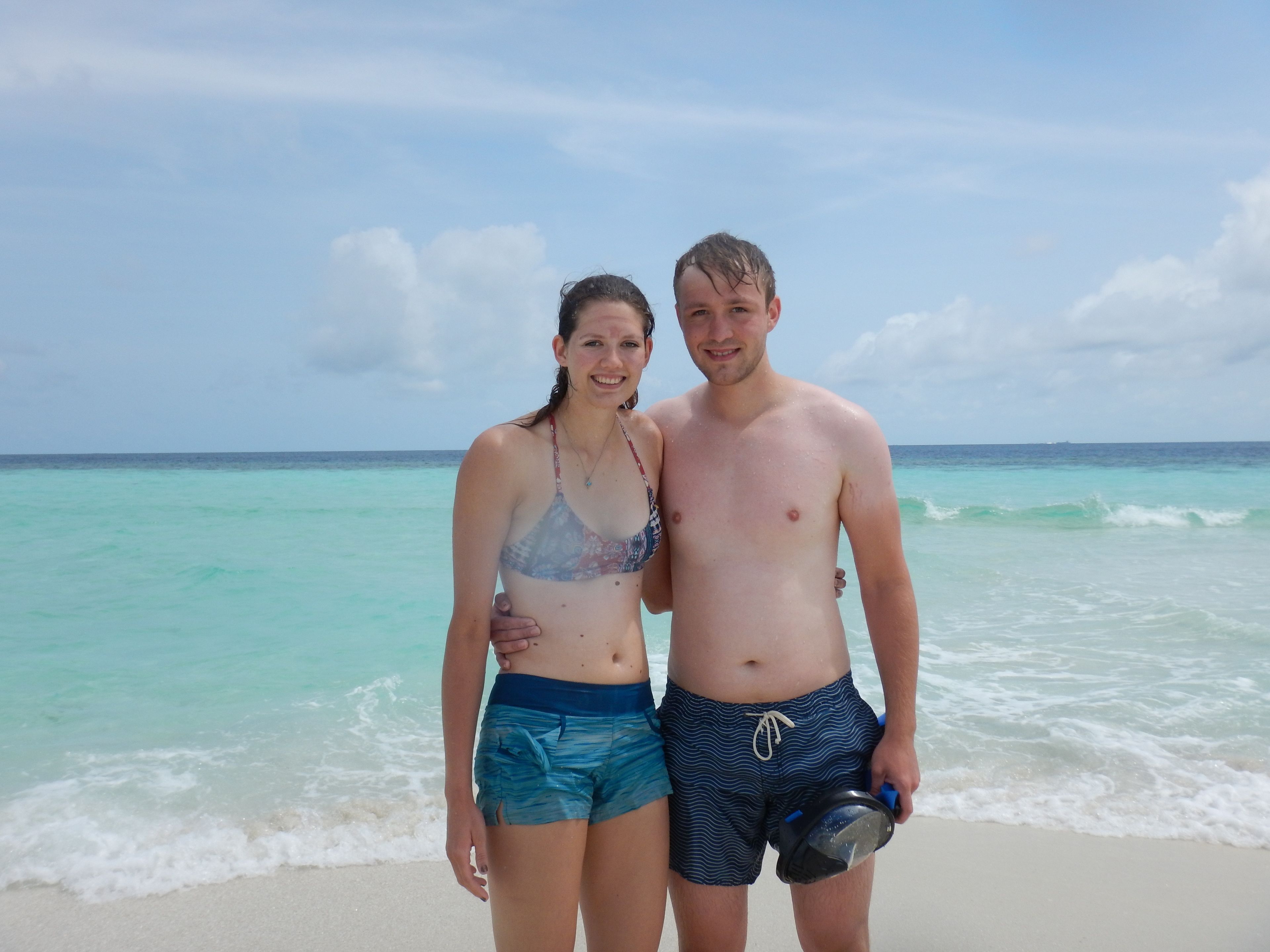 A beach scene with two people standing close together, a clear sky, and turquoise ocean water.