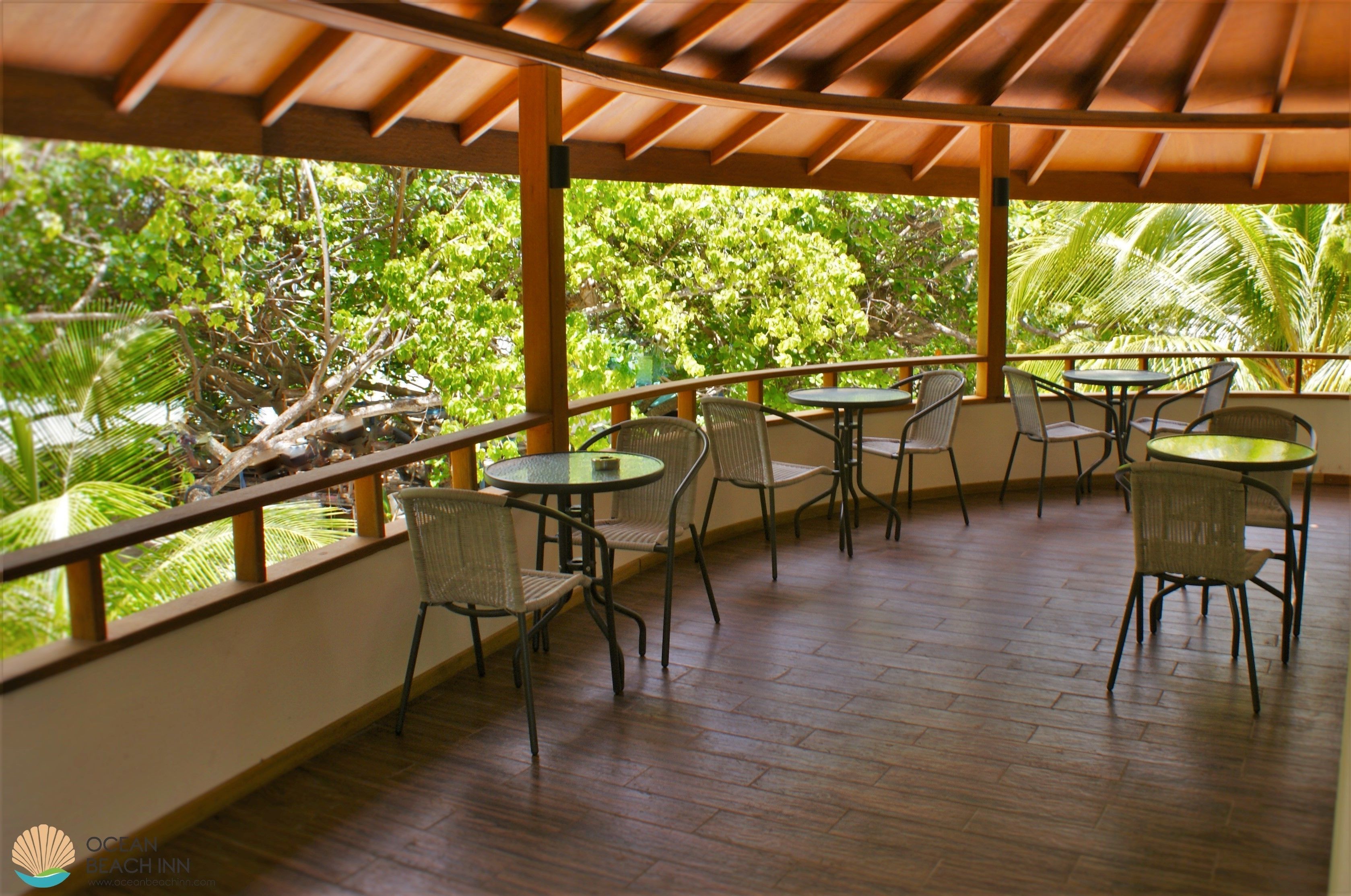 Un espacio al aire libre cubierto, con suelo de madera, sillas y mesas de metal, rodeado de abundante vegetación y palmeras.