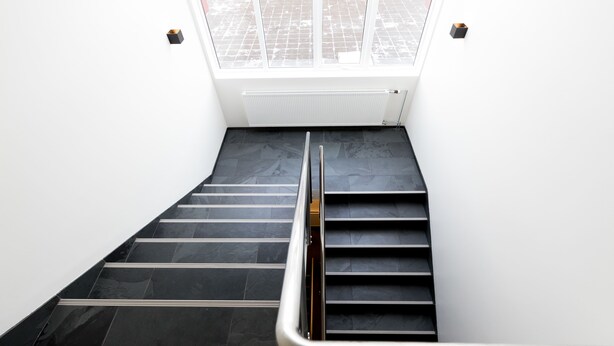 A modern staircase with black tiles and a metal handrail, leading to a bright area with a view of a tiled courtyard. - Hrímland Apartments (Akureyri)