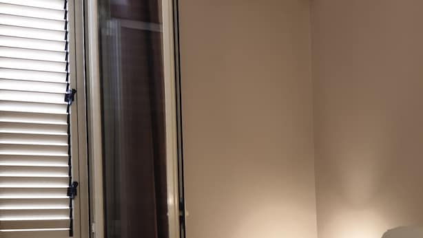 A hotel room with a bed, a nightstand with a lamp, a wall-mounted air conditioner, and a door with blinds. - Oasi Iblea (Ragusa)