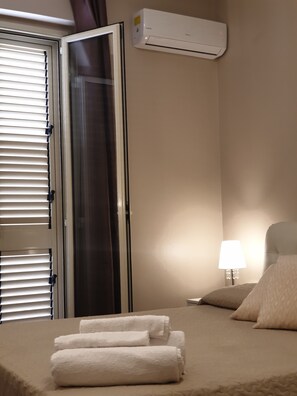 Una habitación de hotel con una cama, una mesita de noche con una lámpara, un aire acondicionado instalado en la pared y una puerta con persianas.