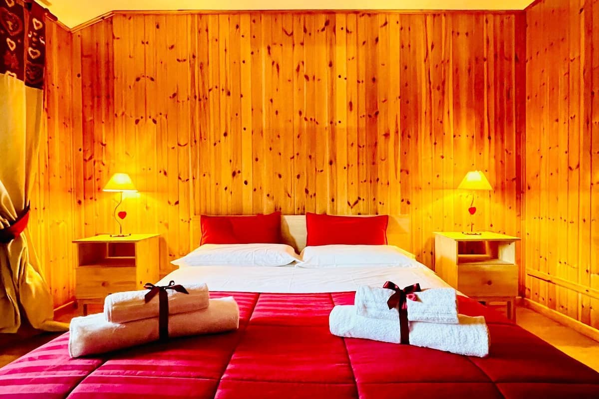 Quarto de hotel com parede de painel de madeira, uma cama grande com roupa de cama vermelha, duas mesas de cabeceira com abajures e um tapete vermelho.