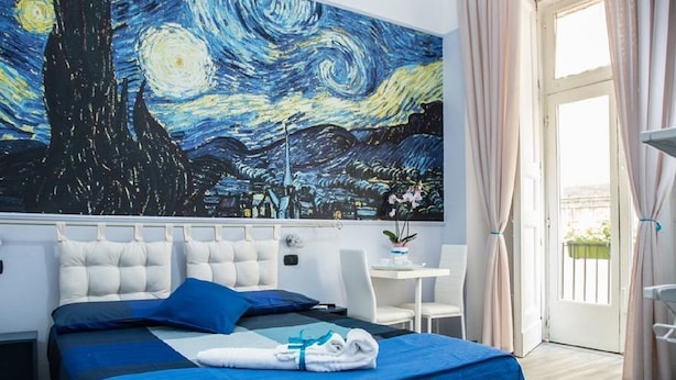 A hotel room with a large painting of a night scene, a bed with blue bedding, a white headboard, and a small table with a chair. - Art In B&B (Salerno)