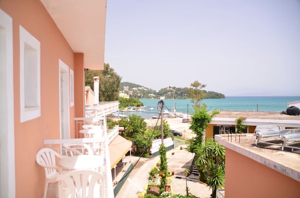 A balcony with white chairs, a view of the sea, and a building with a red facade. - Hotel Sirena Beach (Corfu)