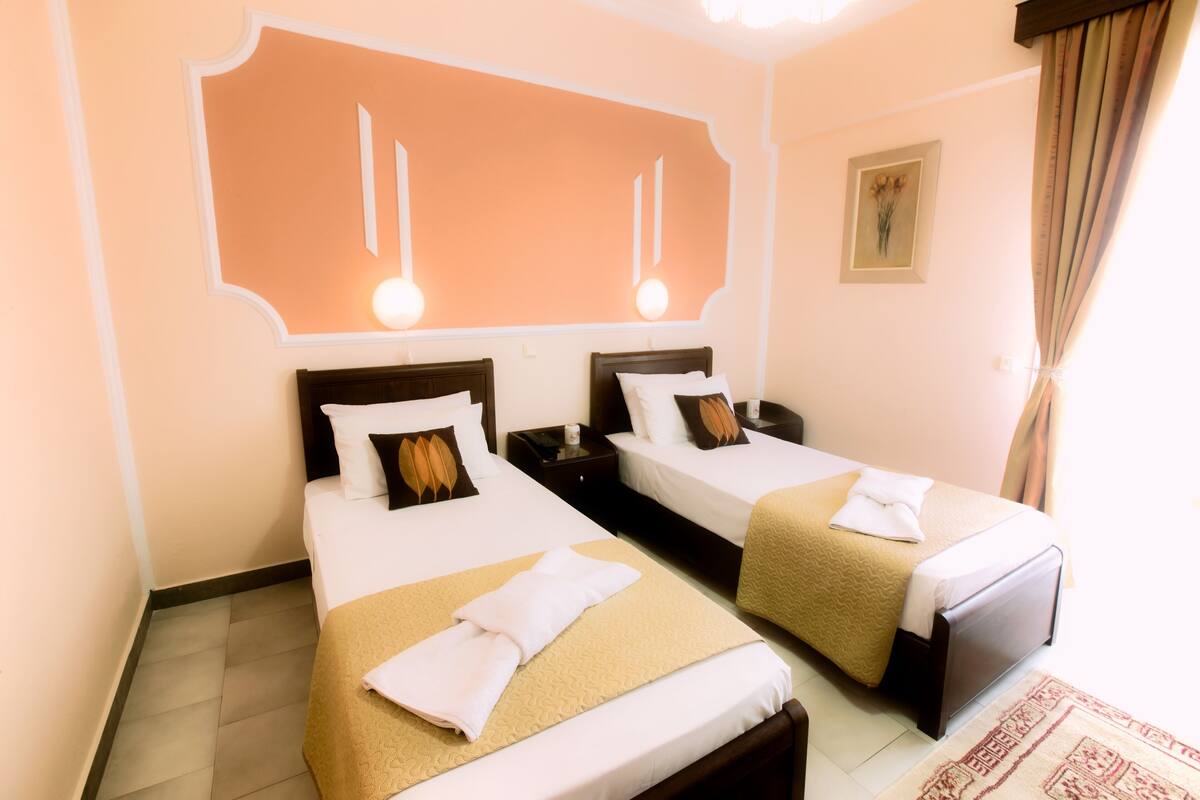 A hotel room with two beds, each with a beige bedspread and white linens, a large mirror above the beds, and a framed picture on the wall.