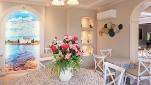 A room with a large window, a white chair, a table with a patterned tablecloth, and a vase of flowers. - Hotel Sirena Beach (Corfu)