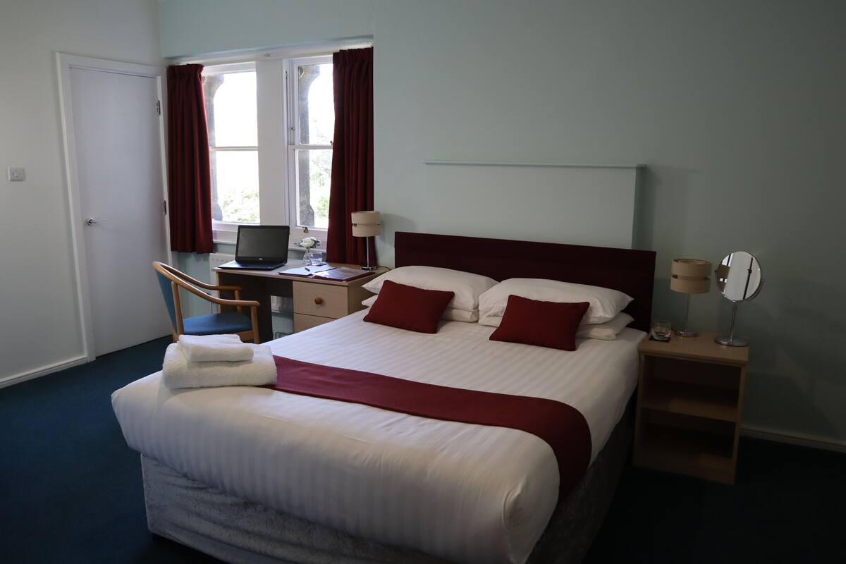 A hotel room with a bed, a desk with a computer, a chair, a bedside table with a lamp, and a window with curtains.
