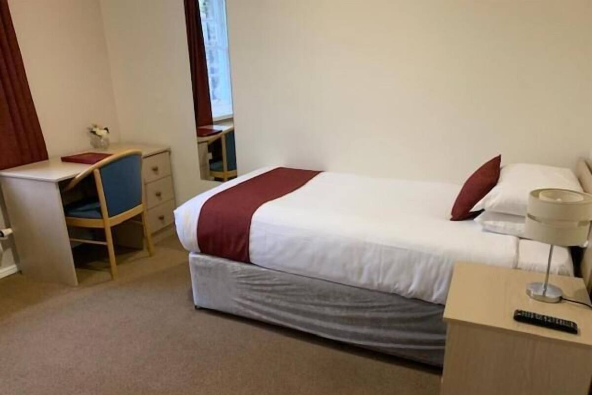 A hotel room with a bed, a desk, a chair, a lamp, and a remote control.