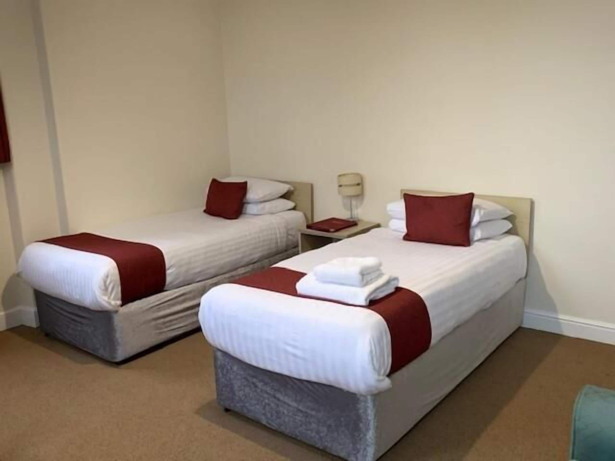 A hotel room with two single beds, each with a red pillow and white linens. There is a nightstand with a lamp between the beds.