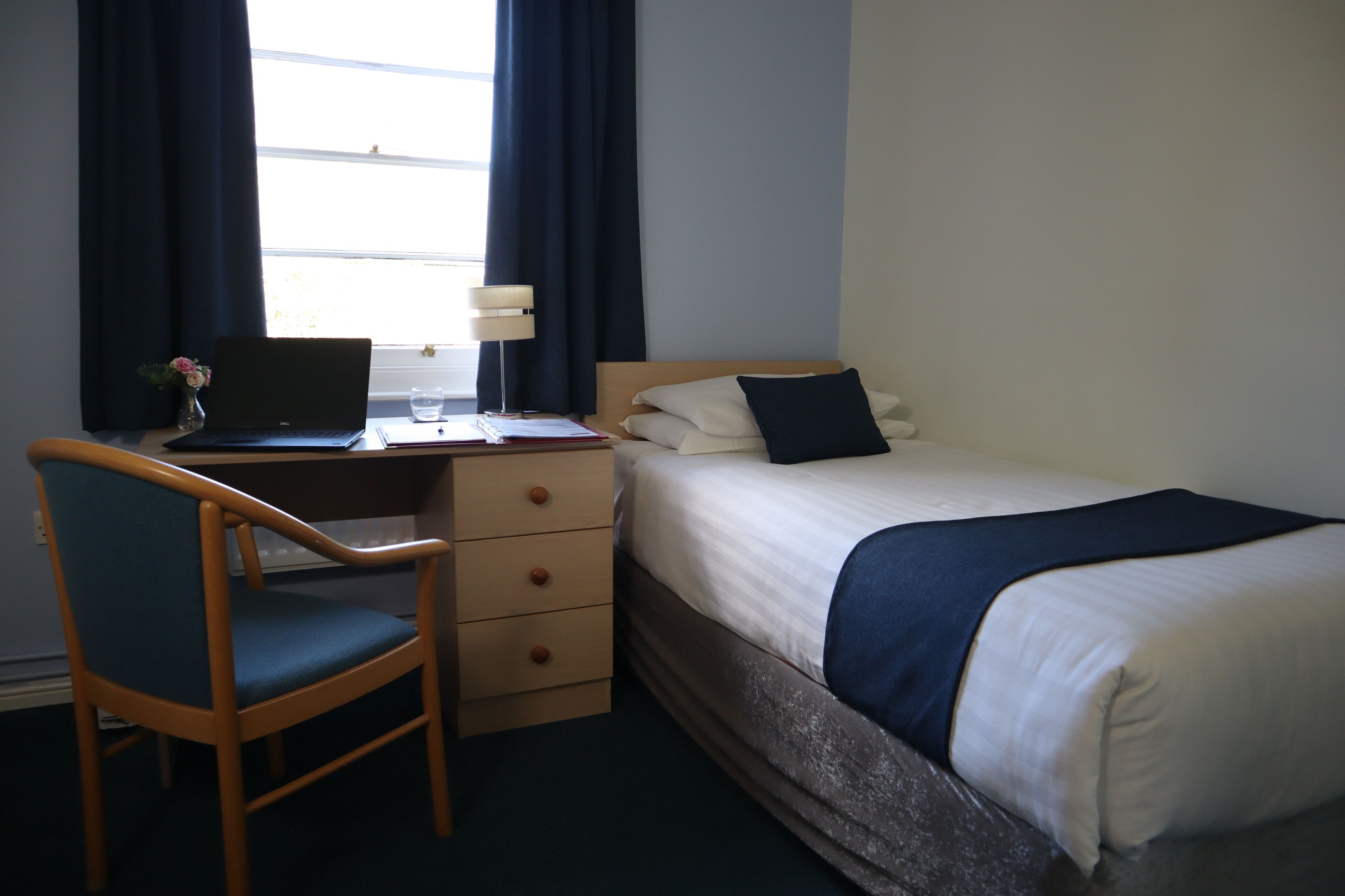 A hotel room with a bed, a desk with a laptop, a chair, and a window with curtains.