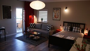 Ein modernes Schlafzimmer mit Bett, Sofa, Couchtisch und Esstisch. Ein Fenster mit Jalousien, ein roter Vorhang und ein Poster an der Wand.