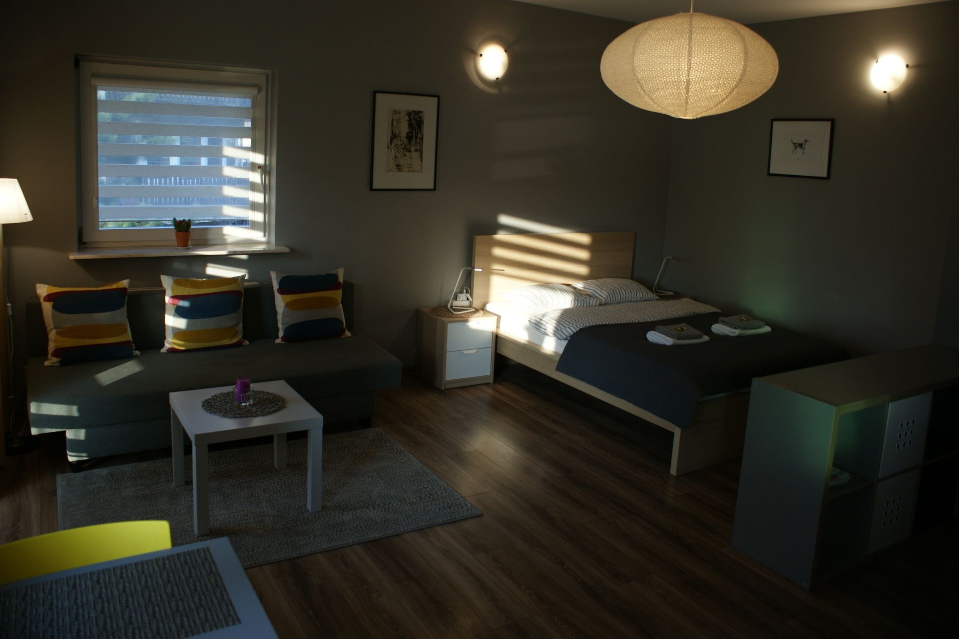 A modern bedroom with a bed, bedside table, lamp, and a window with blinds.