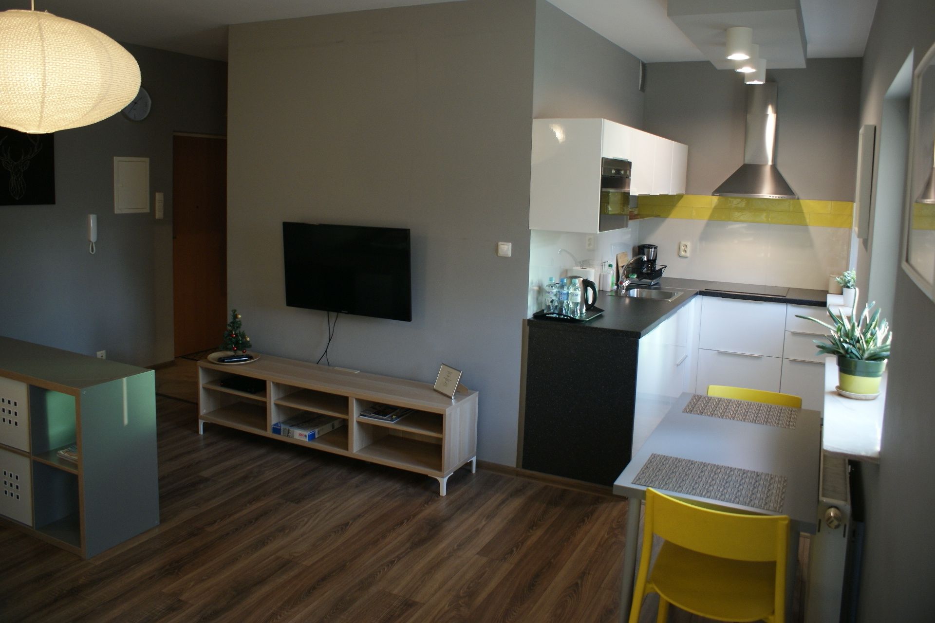 Un espacio habitable compacto con kitchenette, un comedor con sillas amarillas, un televisor montado en la pared y un pequeño árbol de Navidad.