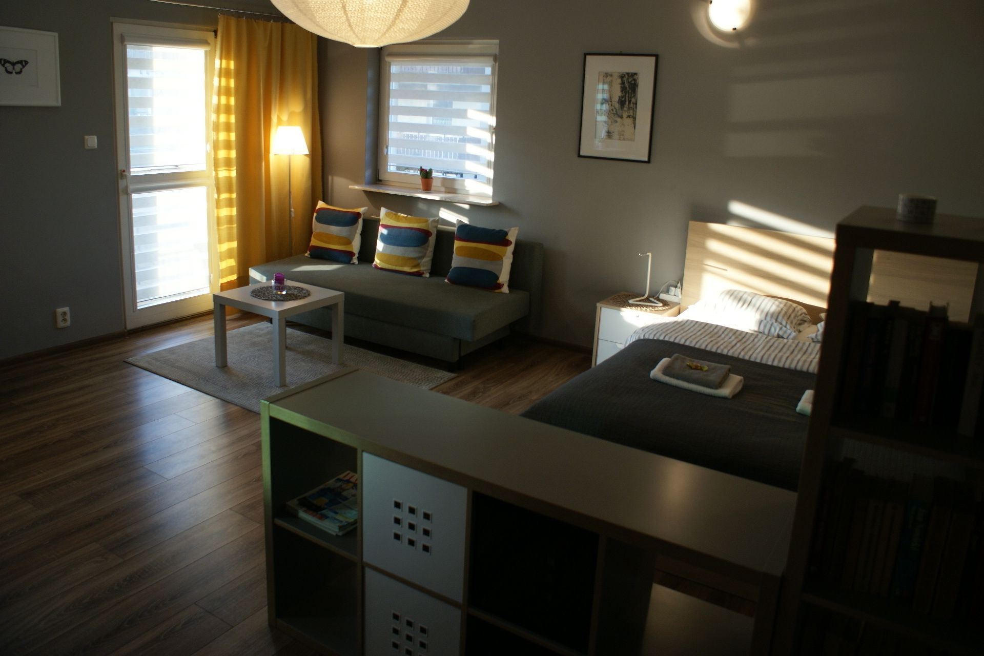 A modern living room with a sofa, a small table, and a bed. There are windows with blinds, a lamp, and a picture on the wall.