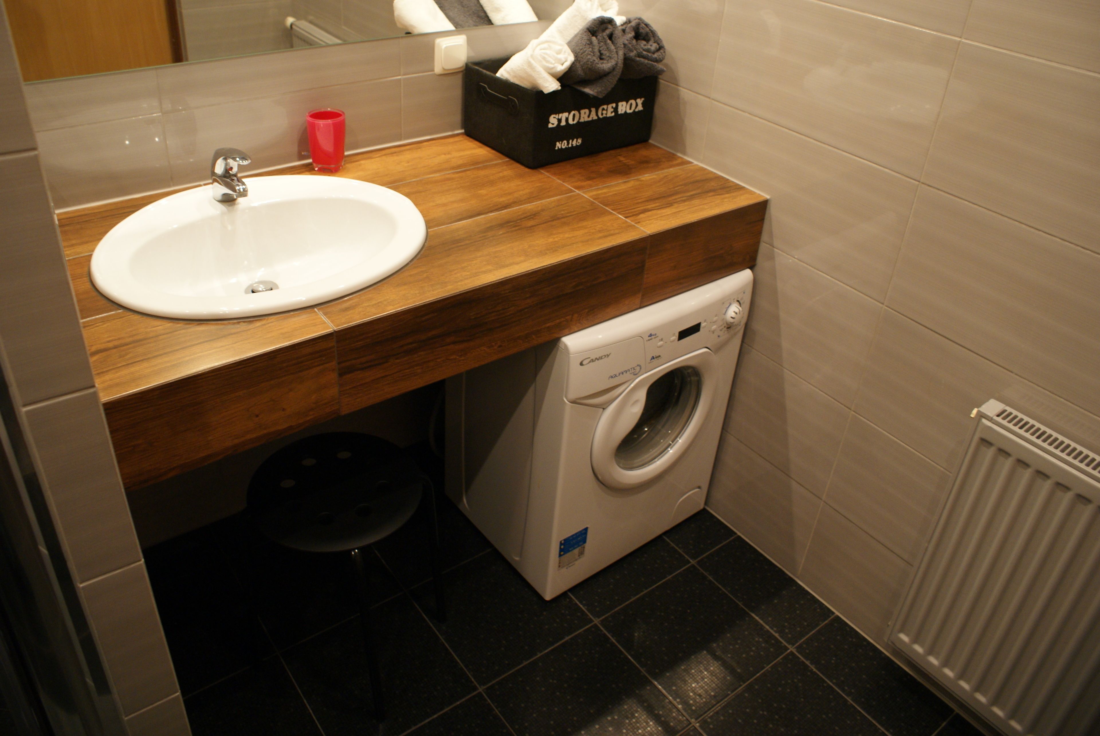 Ein kleines Badezimmer mit einem hölzernen Waschtisch, einem weißen Waschbecken, einer Waschmaschine und einer Aufbewahrungsbox mit der Aufschrift 'STORAGE BOX'.