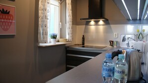 Eine moderne Küche mit eingebautem Herd, einem Fenster mit gemusterten Vorhängen und einer Arbeitsplatte mit Wasserflaschen und einem Wasserkocher.