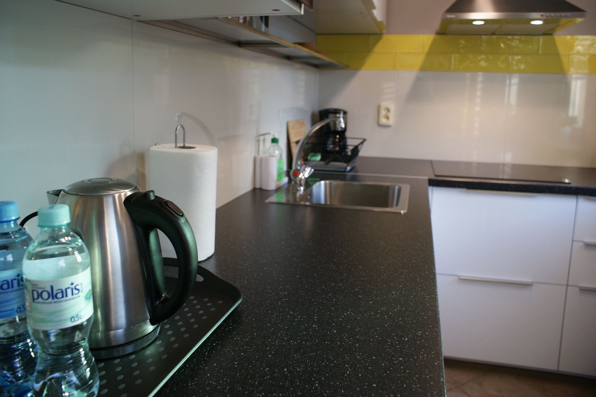 A kitchen with a black countertop, a kettle, two water bottles, a roll of paper towels, and a coffee maker.