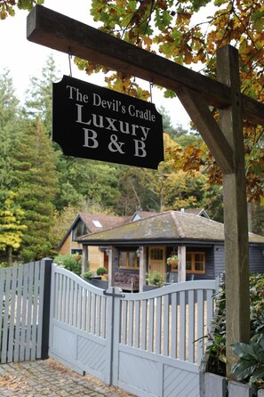 Een bordje voor 'The Devil's Cradle Luxury B&B' met een houten poort en een huis op de achtergrond.
