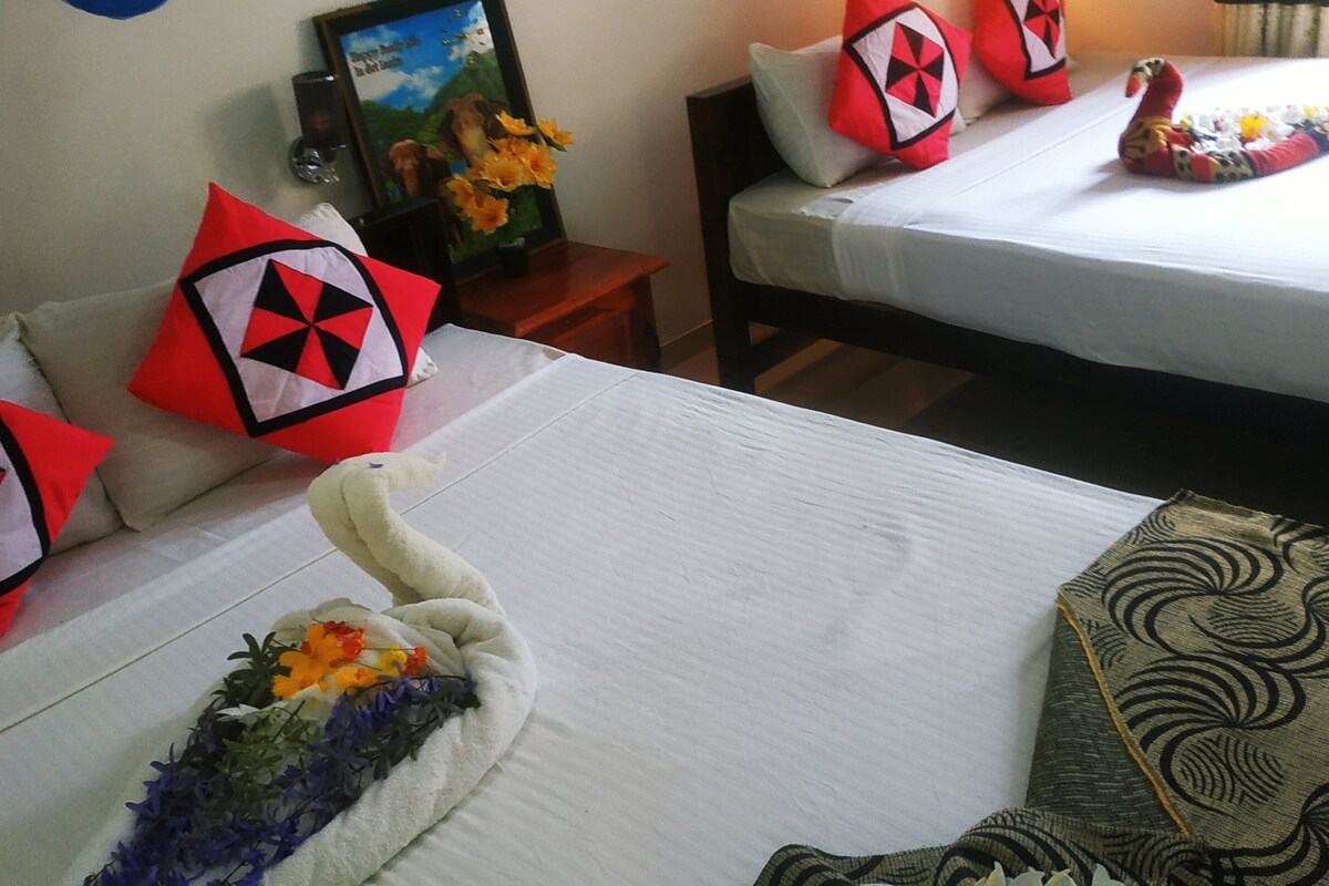 A hotel room with two beds, each with a white bedspread and red pillows with a black geometric pattern. There is a framed picture on the wall and a small table with a vase of flowers.