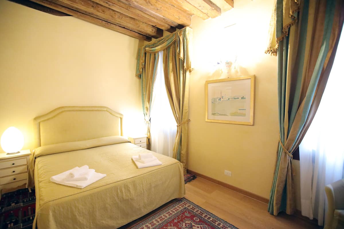 Habitación de hotel con cama, mesita de noche, lámpara, cortinas, un cuadro enmarcado en la pared y techo de madera.