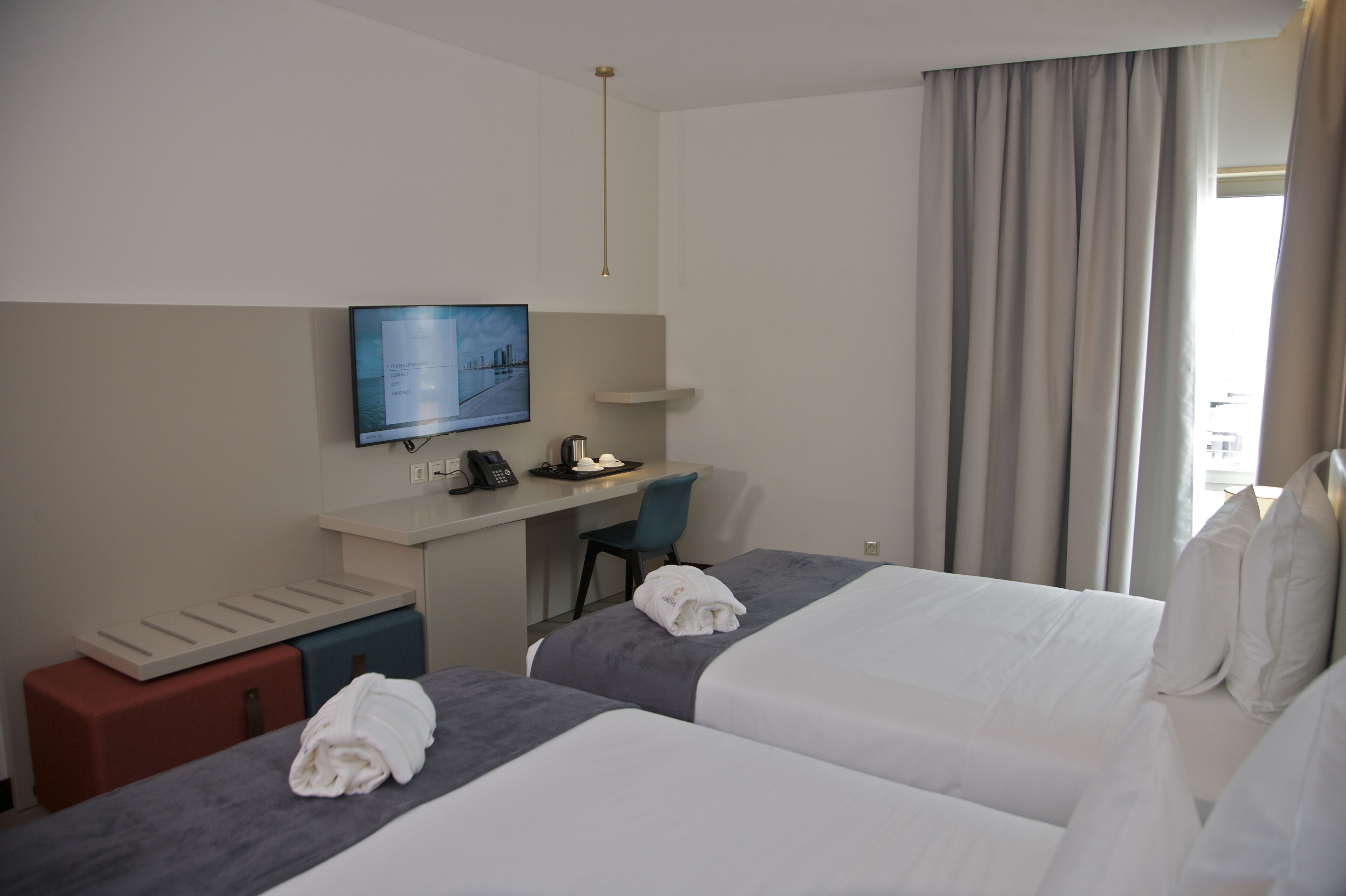 Et hotelværelse med to senge, et skrivebord med en computer, et fjernsyn og et vindue med gardiner.