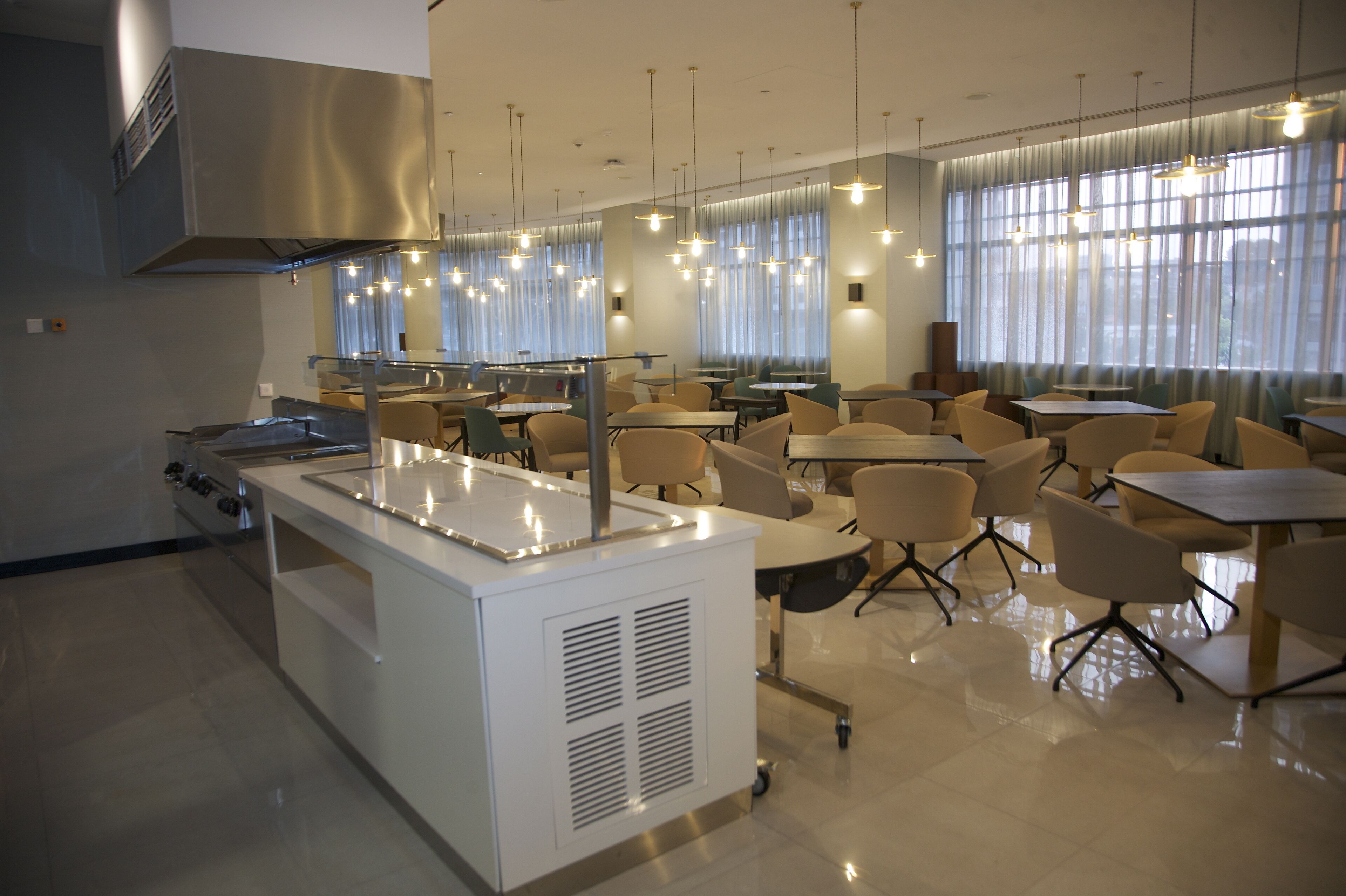 A modern dining area with a central kitchen station, multiple tables and chairs, and large windows with curtains.