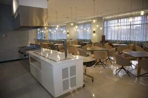 A modern dining area with a central kitchen station, multiple tables and chairs, and large windows with curtains.