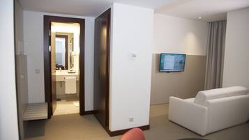 Habitación de hotel con sofá, televisión, baño con lavamanos y espejo, y una puerta que da a otra habitación.