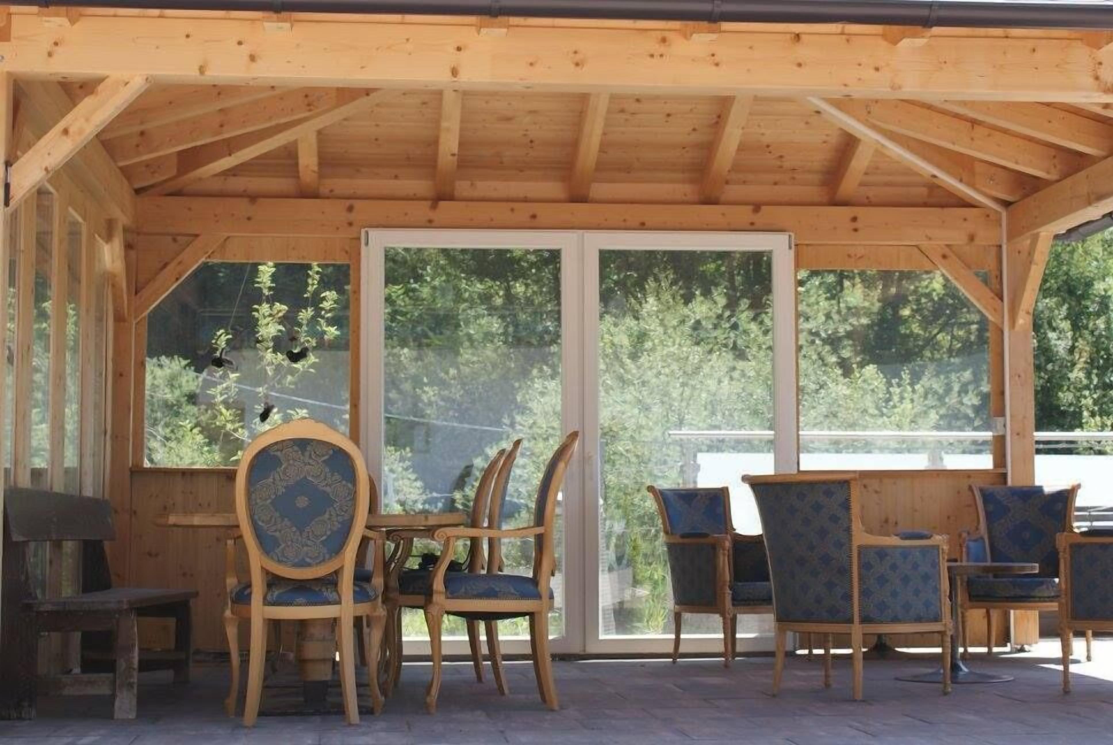 A wooden covered patio with a table and chairs, large windows, and a view of trees.