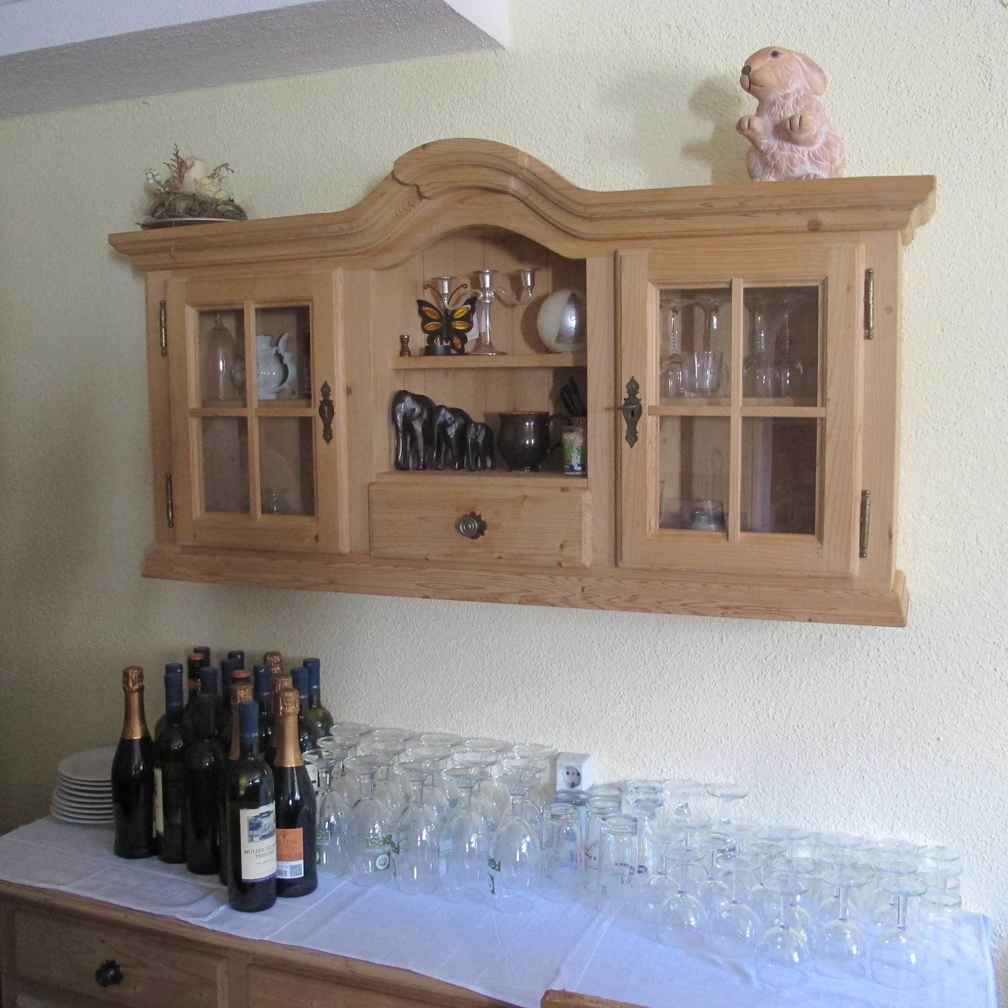 Un buffet en bois doté de portes en verre, renfermant divers objets en verre et éléments décoratifs. En dessous, on trouve une table sur laquelle sont disposées des bouteilles de vin et des verres.