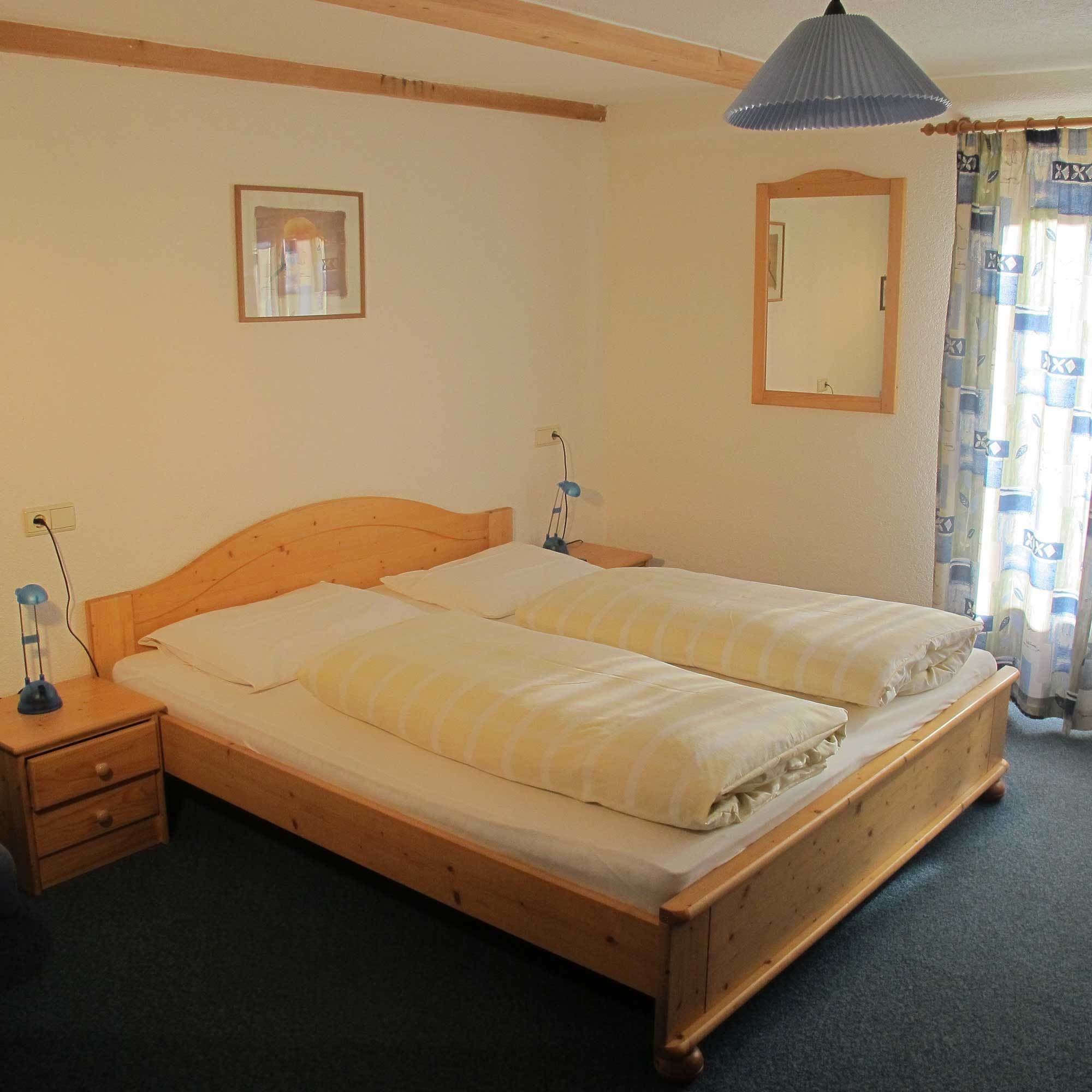 A single-bed room with a wooden headboard, a bedside table with a lamp, a mirror, and a window with curtains.