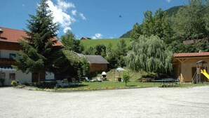 A rural setting with a gravel parking area, a wooden building with a red roof, and a playground with a slide. - Pension Wirt am Bach (Terento)