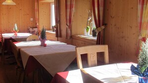 A wooden interior with tables and chairs, a hanging light fixture, and a window with curtains. - Pension Wirt am Bach (Terento)