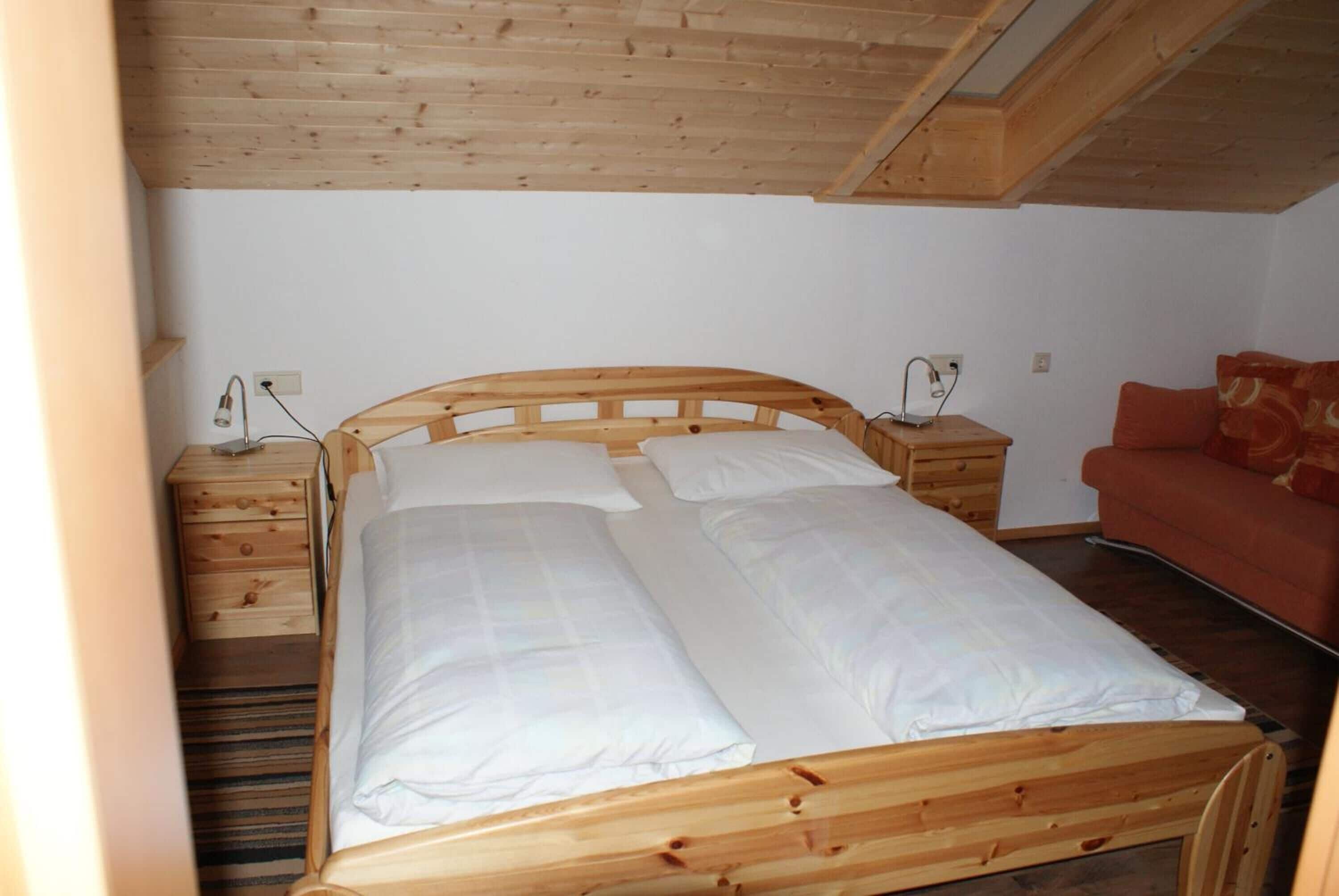 A wooden bed with white linens in a room with a sloped ceiling and a red sofa.