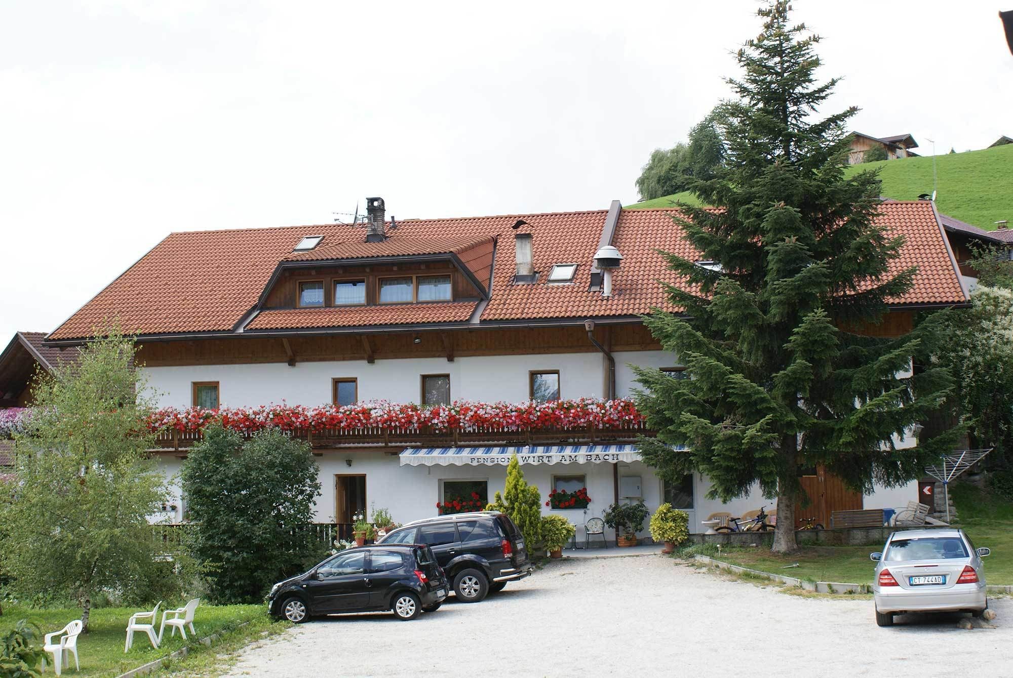 A traditional chalet-style building with a red-tiled roof, a balcony with red flowers, and a parked car in front.
