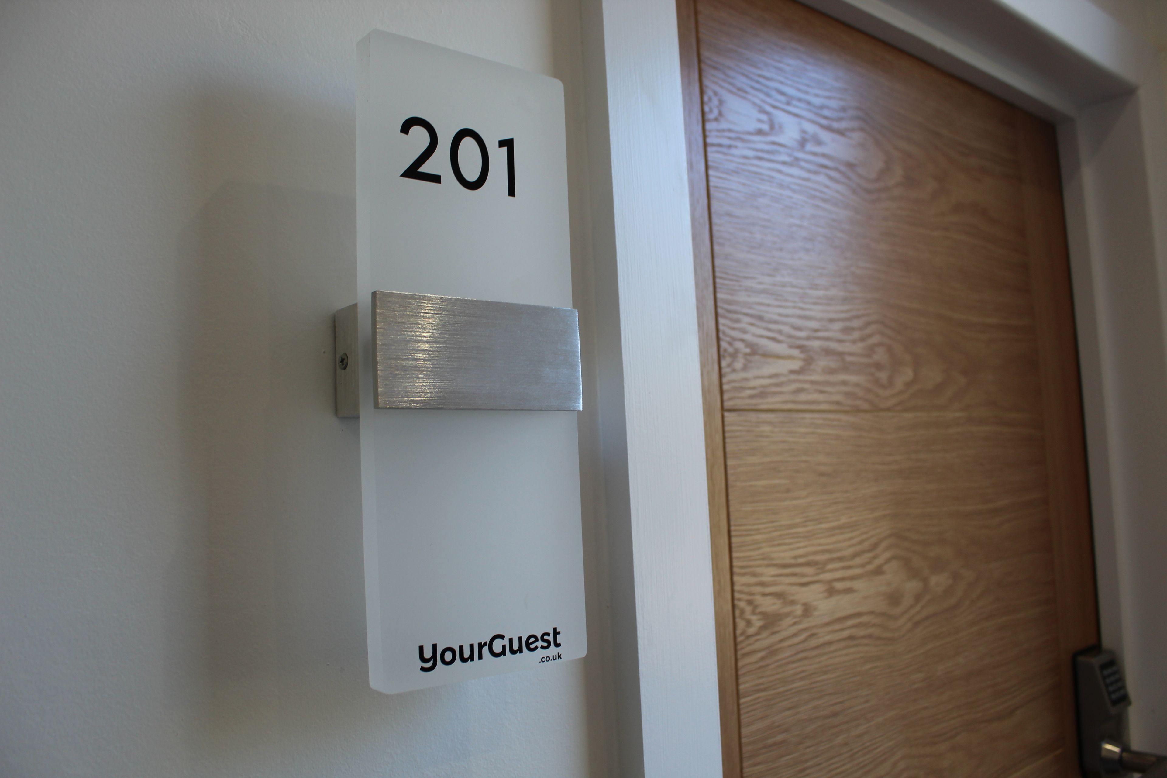 Une plaque indiquant le numéro de chambre, portant le numéro 201 et le texte « YourGuest.co.uk », est fixée à un mur blanc.