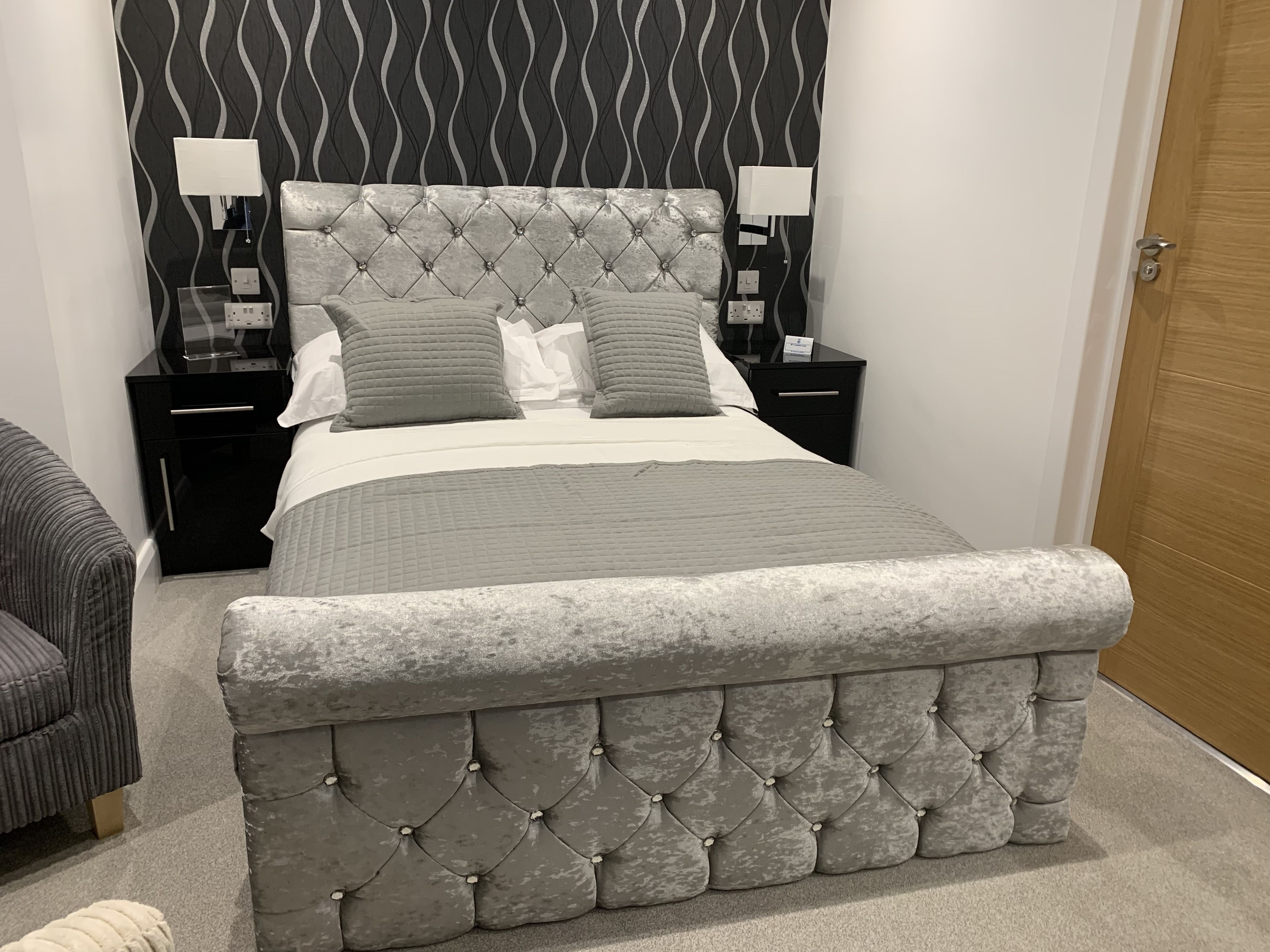 A bedroom with a tufted headboard, a bed with grey bedding, two bedside tables with lamps, and a wooden door.