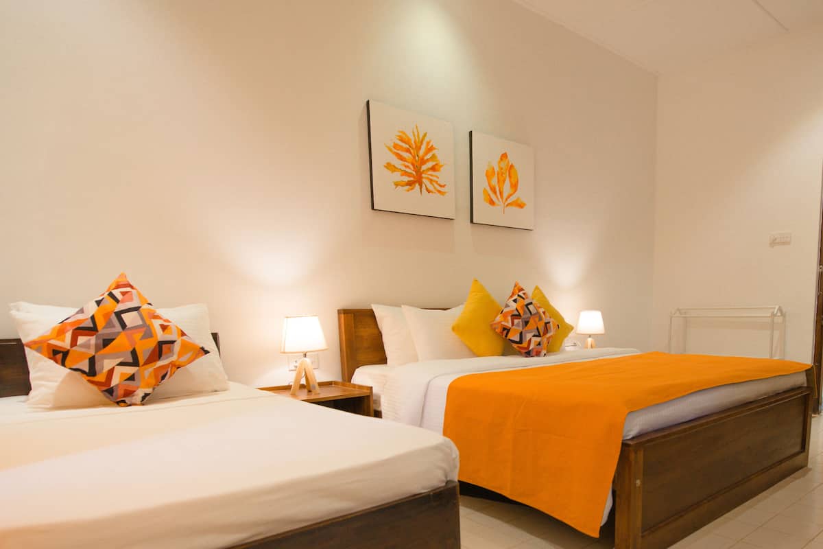 Ein Hotelzimmer mit zwei Betten, beide mit weißer Bettdecke und Kissen, und eine orangefarbene Tagesdecke auf einem Bett. Es gibt zwei Nachttische mit Lampen, und die Wände sind mit zwei gerahmten Blattform-Kunstwerken geschmückt.