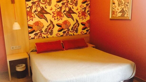 A bedroom with a bed, red pillows, a nightstand, a lamp, and a floral headboard. - Hotel Ginebra (Barcelona)