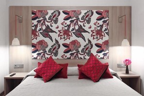 A hotel room with a bed, two red pillows, a nightstand, a lamp, and a wall with a floral pattern.