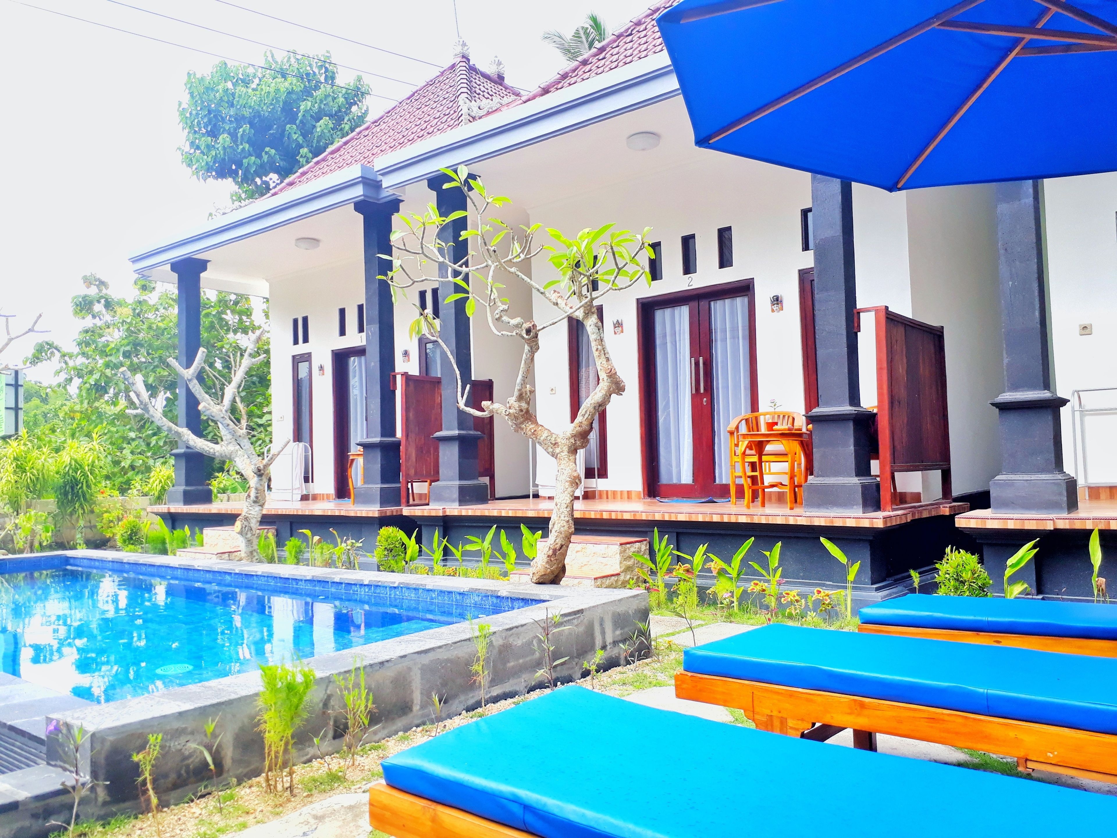 Un espace aménagé au bord de la piscine, avec un parasol bleu, des chaises longues en bois et un bâtiment doté de piliers sombres et de portes rouges.