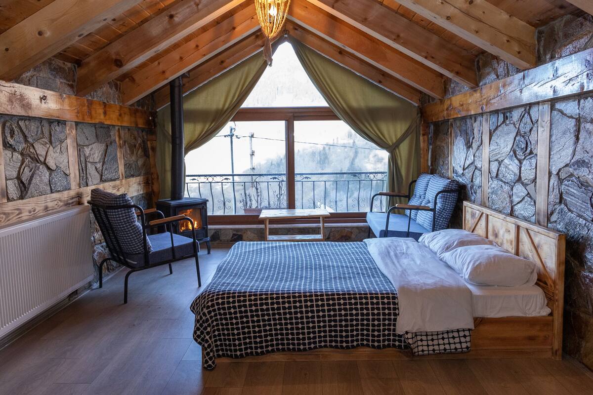 A cozy attic bedroom with a bed, two chairs, a small table, and a view of the outside.