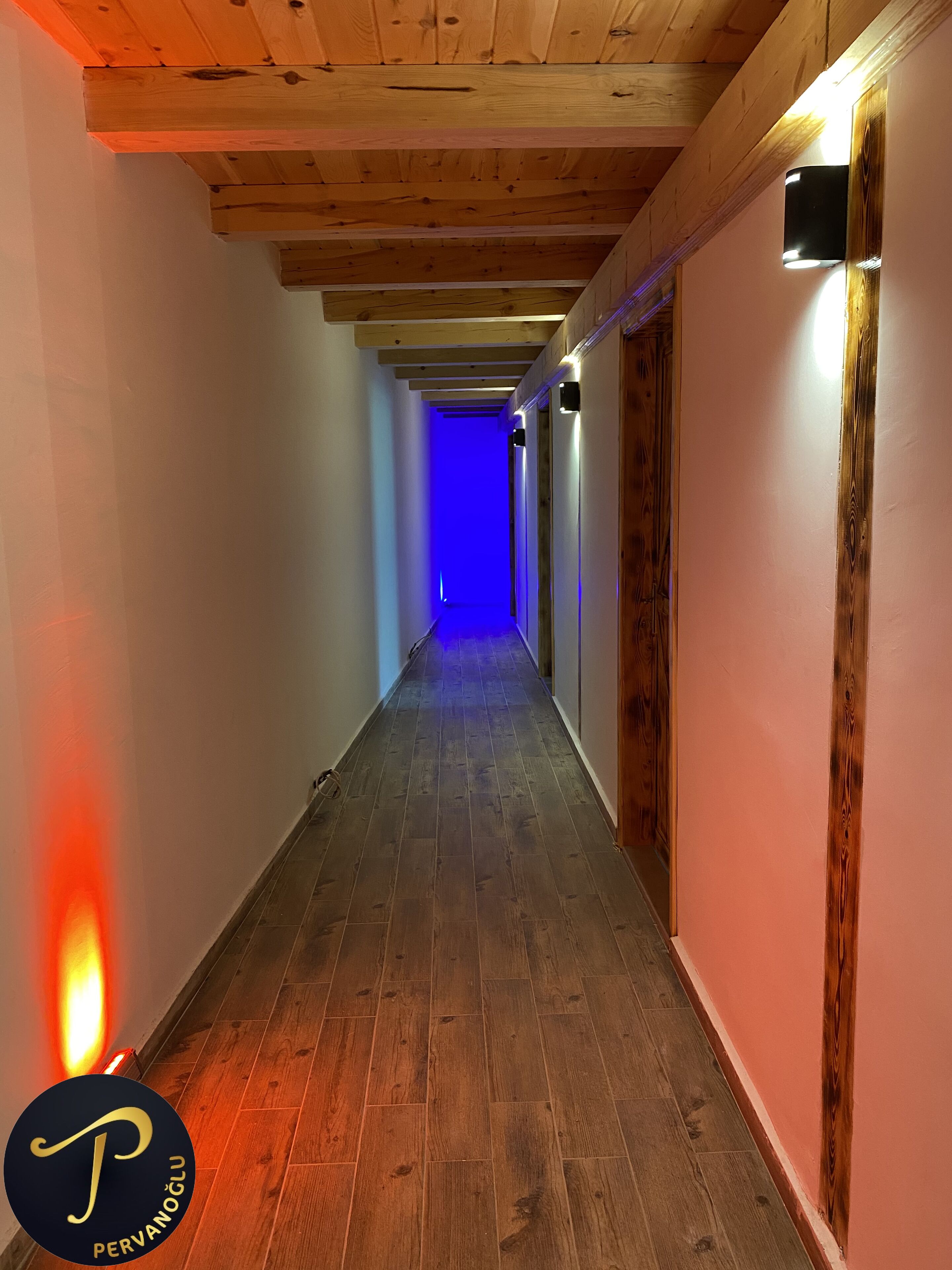 A narrow hallway with wooden flooring and exposed beams, leading to a room with a blue-lit wall.