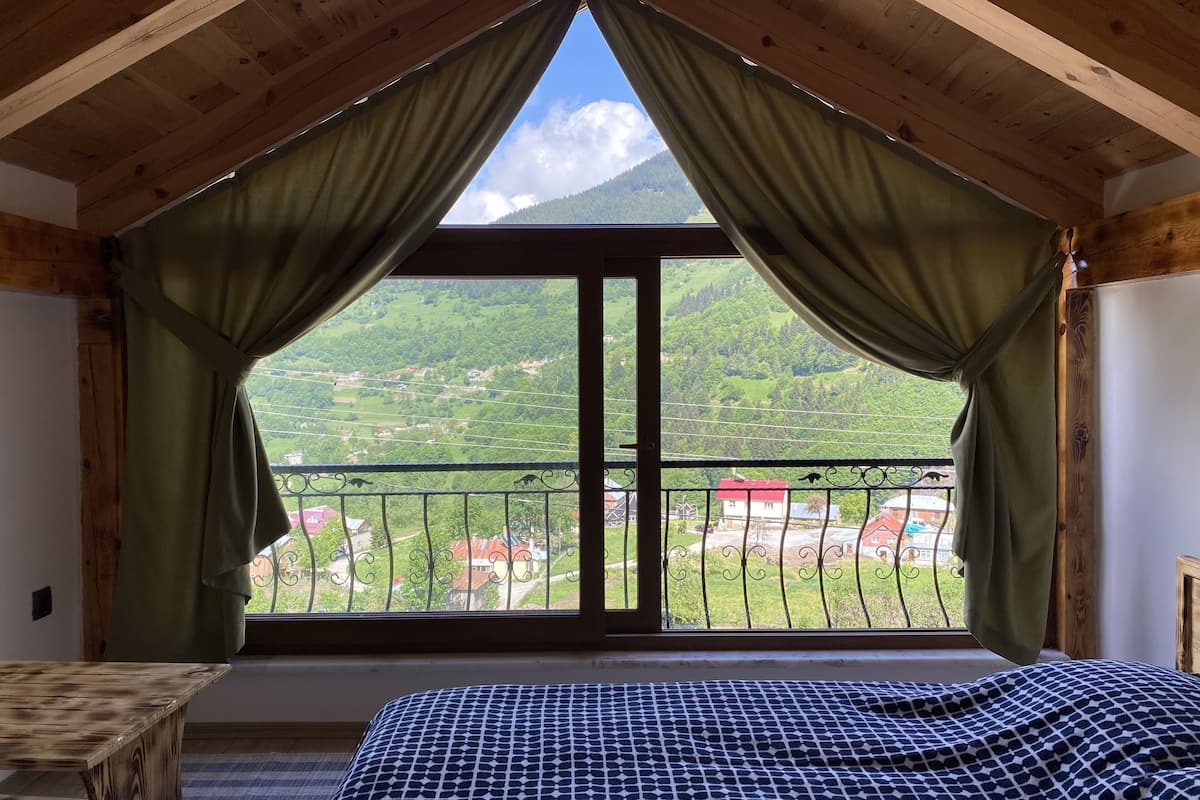 A room with a large window, wooden ceiling beams, a bed with a patterned bedspread, and a view of a green landscape with houses.