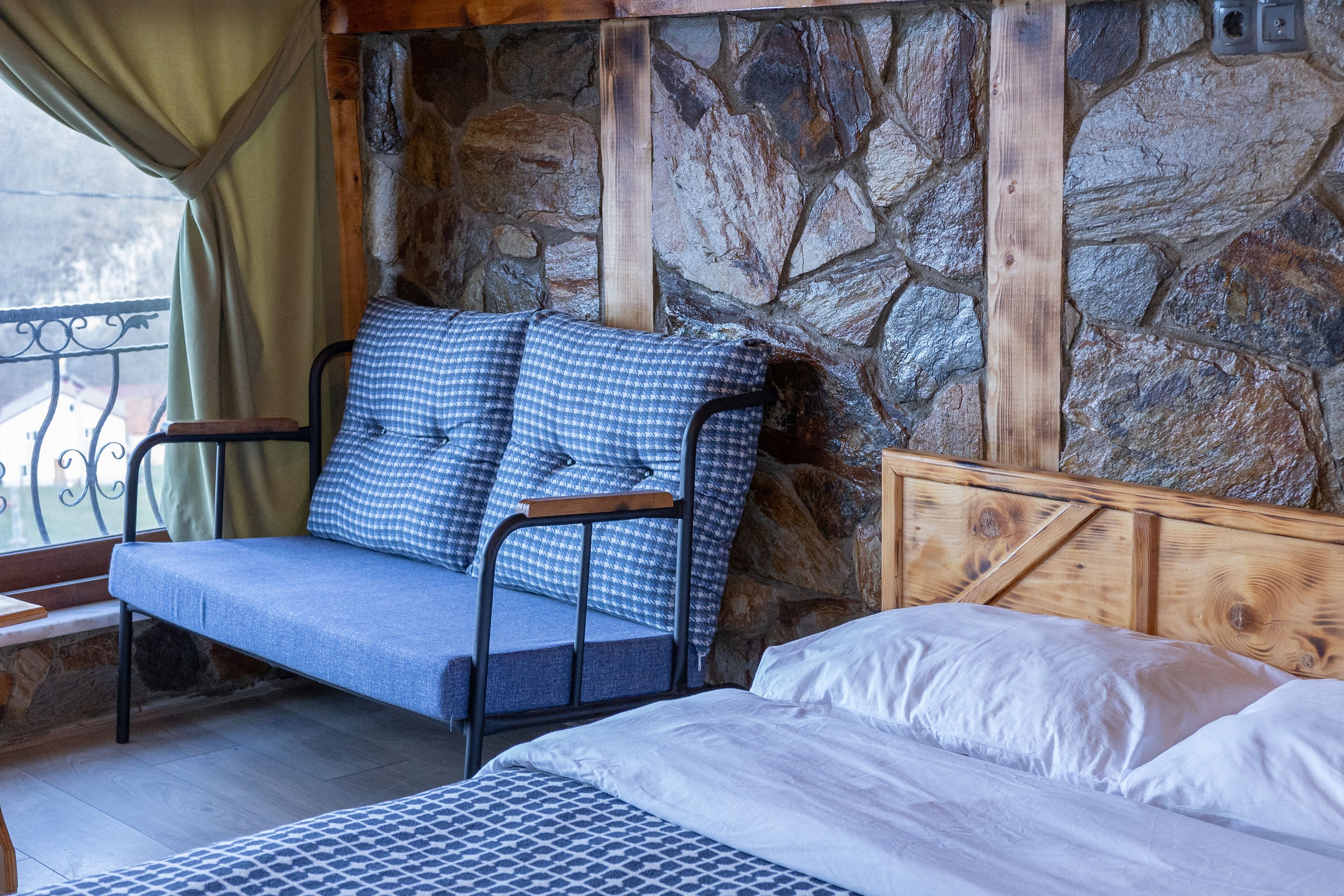 A cozy room with a stone wall, a wooden headboard, a checkered sofa, and a patterned bedspread.