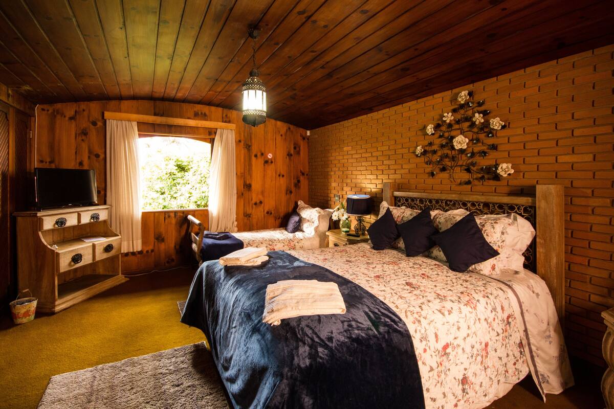Quarto de cabana de madeira com duas camas, uma televisão, uma cômoda e uma parede de tijolos decorada com motivos florais.