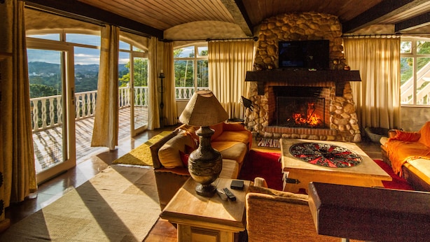 A cozy living room with a stone fireplace, comfortable seating, and a large window overlooking a scenic landscape. - Pousada Déjà Vu (Campos do Jordão)