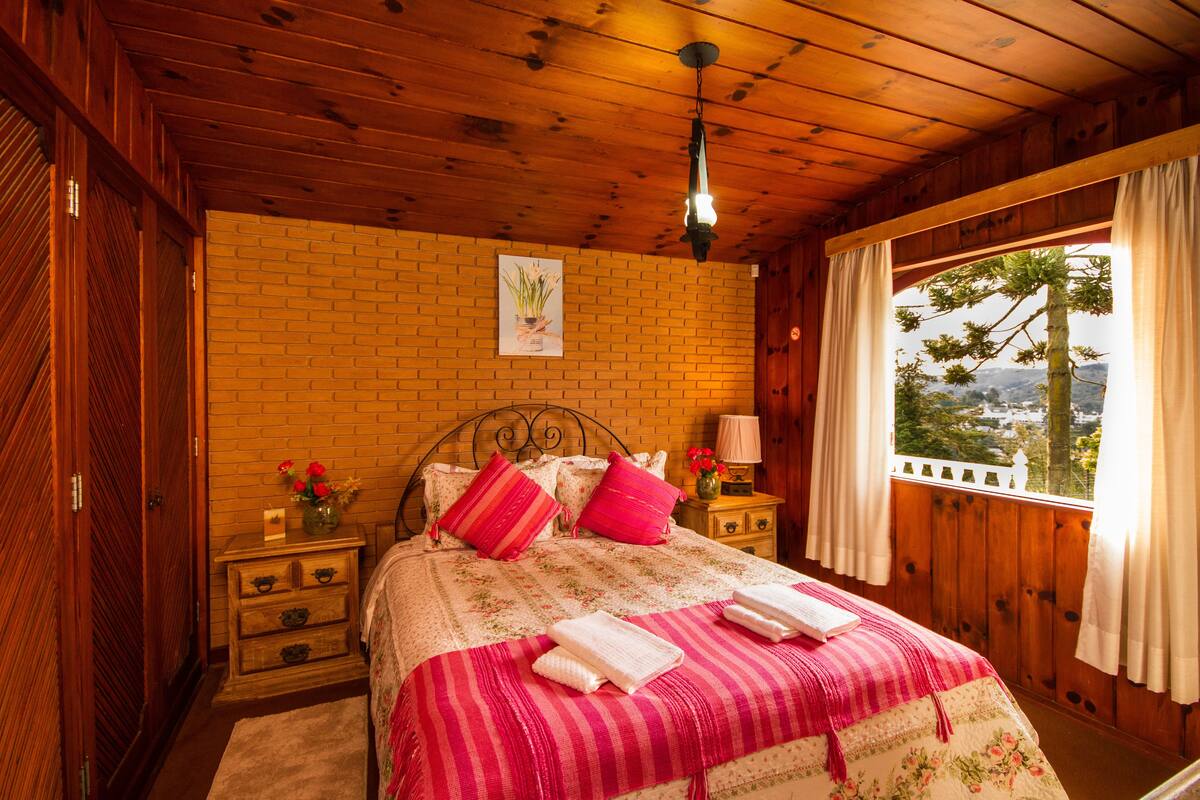 Quarto de cabana de madeira com parede de tijolos, cama com roupa de cama rosa e branca, mesa de cabeceira com abajur e janela com cortinas.